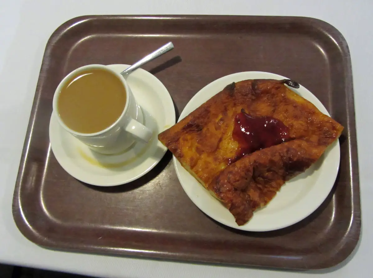 Firkantet stykke gyllenbrun ovnspannekake med rødt syltetøy, servert med en kopp kaffe på et brett