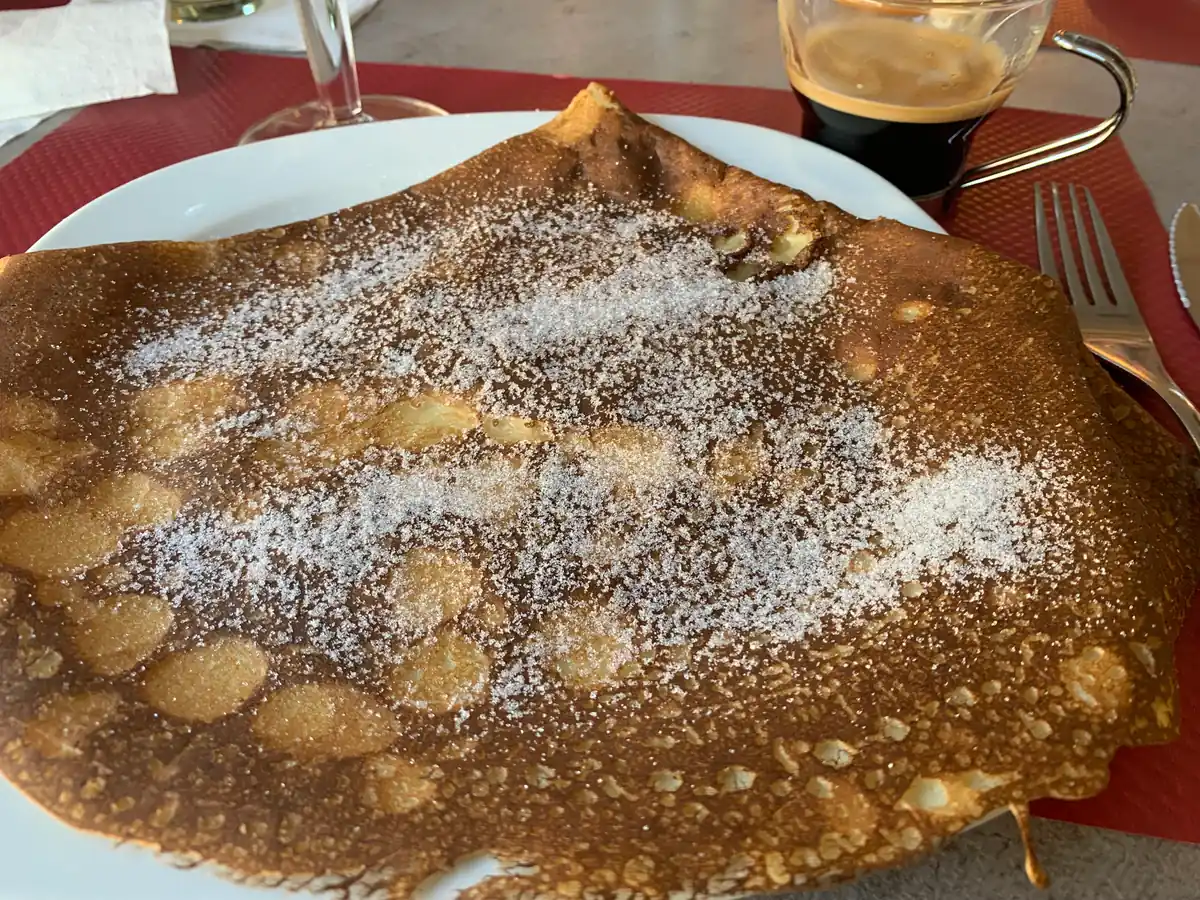 Stor tynn crêpe med strøsukker på en hvit tallerken, med en kopp kaffe ved siden av